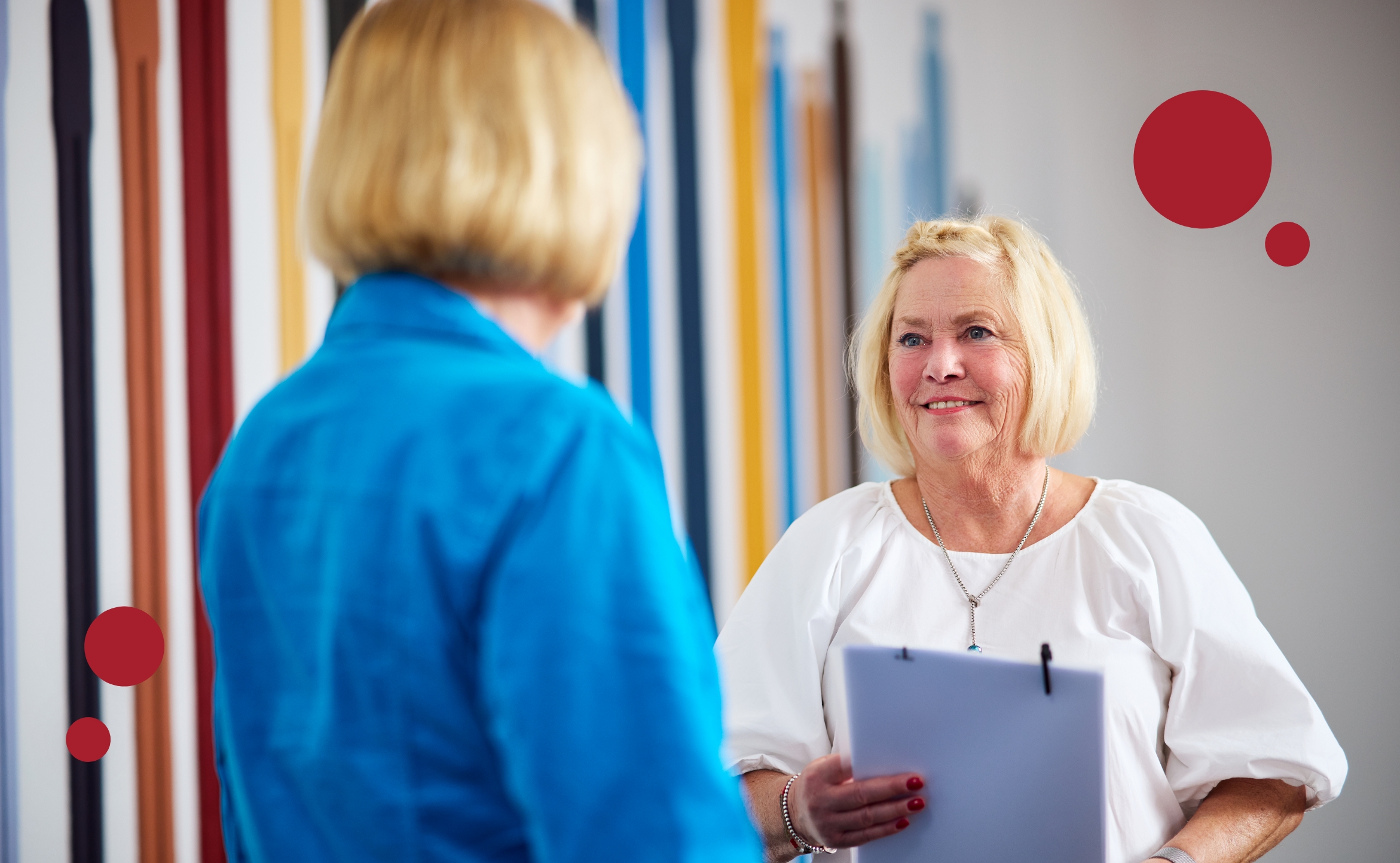 Zwei Frauen mit blonden Haaren stehen sich gegenüber und sprechen, eine hält ein Notizblatt in der Hand.